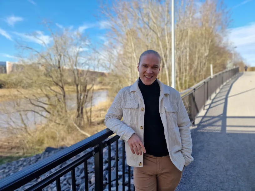 Mies seisoo sillalla aurinkoisena päivänä, puut ja joki taustalla.