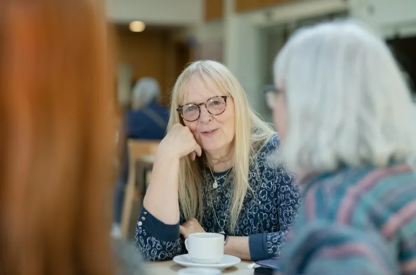 Kolme naista keskustelee kahvilassa, keskellä nainen hymyilee ja kuuntelee.