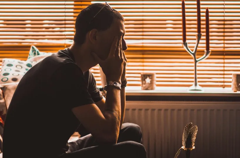 Mies istuu sohvalla, pää käsissään, valo siivilöityy sälekaihtimien läpi.