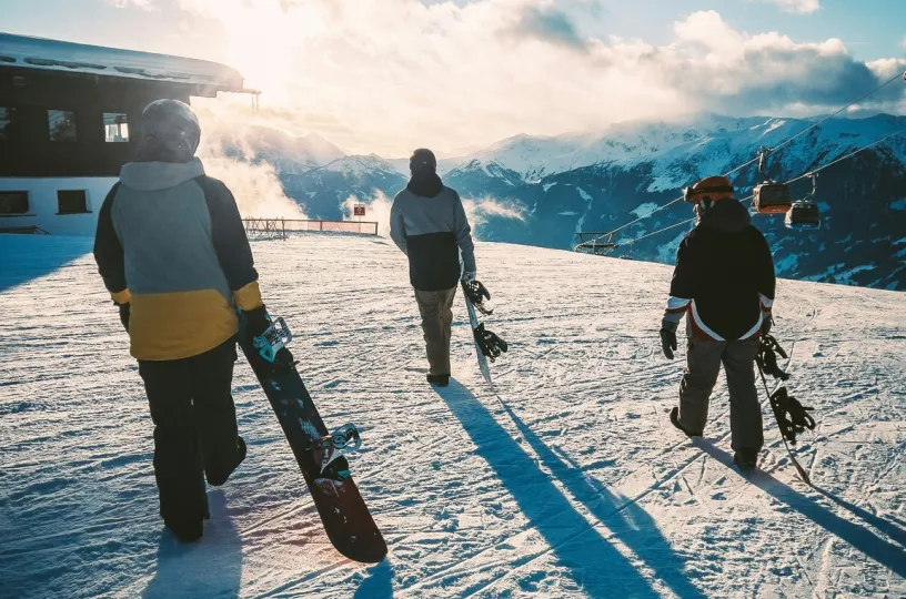 Kolme lumilautailijaa kävelee auringonlaskussa lumisella rinteellä.