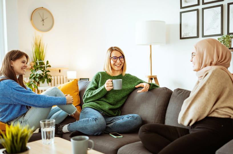 kolme nuorta naista juttelee ja juovat kahvia sohvalla istuen