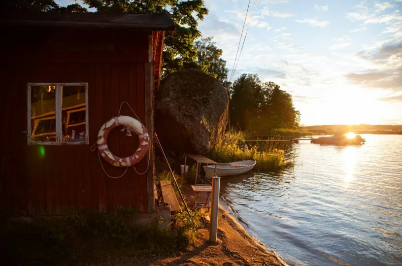 Punainen mökki järven rannalla, vene vedessä ja aurinko laskee taustalla.