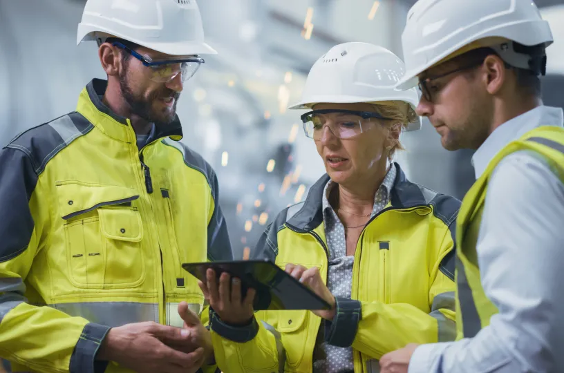 Kolme insinööriä seisoo putkitehtaassa, keskustelevat ja katsovat tablettia. 