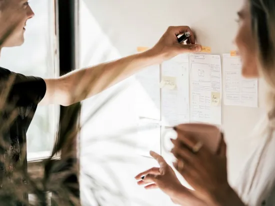 Kaksi henkilöä suunnittelee muistiinpanolappujen äärellä, toinen pitelee kuppia.