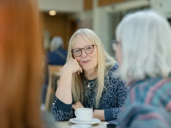 Kolme naista keskustelee kahvilassa, keskellä nainen hymyilee ja kuuntelee.