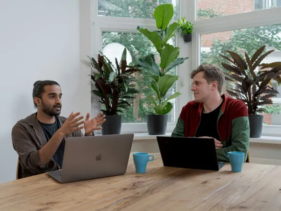 Kaksi henkilöä keskustelee työpöydän ääressä kannettavien tietokoneiden kanssa.