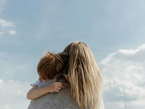 Aikuinen ja lapsi halaavat ulkona sinisen taivaan alla. Kuva on otettu takaapäin, aikuisesta näkyy takaraivo ja lapsen pää aikuisen olalla. 