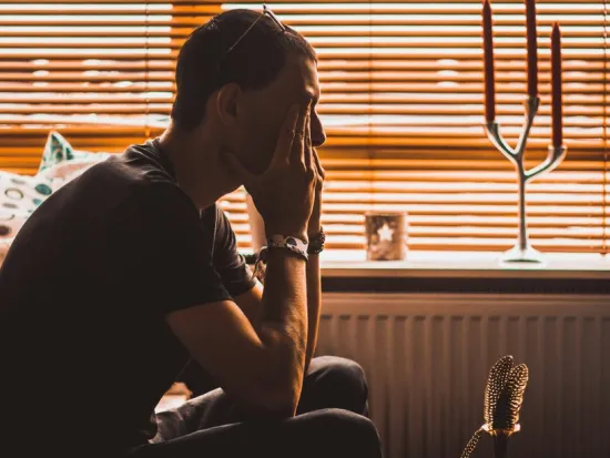 Mies istuu sohvalla, pää käsissään, valo siivilöityy sälekaihtimien läpi.