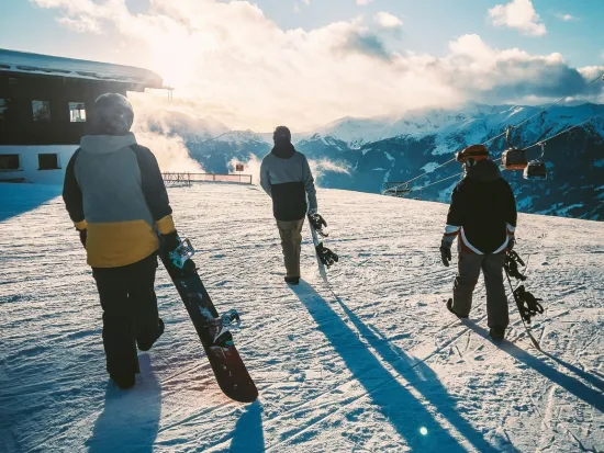 Kolme lumilautailijaa kävelee auringonlaskussa lumisella rinteellä.