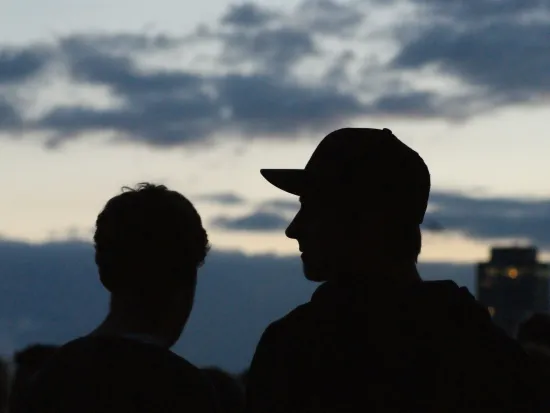 Kaksi henkilöä siluettina iltataivasta vasten, taustalla pilviä ja rakennus.