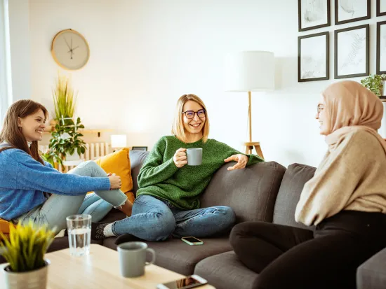 kolme nuorta naista juttelee ja juovat kahvia sohvalla istuen