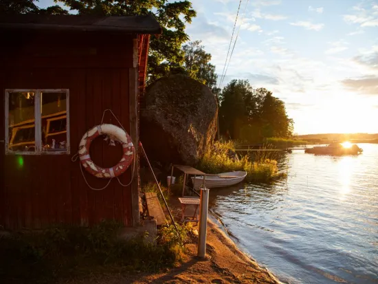 Punainen mökki järven rannalla, vene vedessä ja aurinko laskee taustalla.