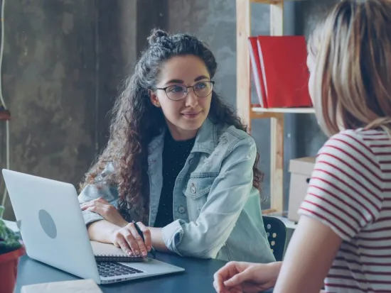 Kaksi naista keskustelee pöydän äärellä, kannettava tietokone edessä.