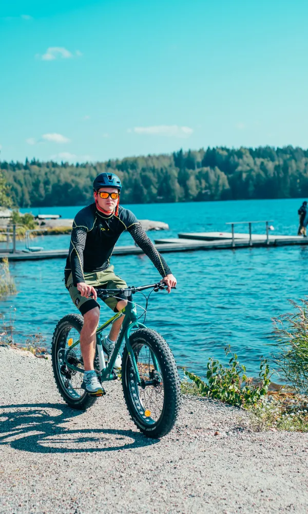 Kaksi pyöräilijää järven rannalla, taustalla ihmisiä melomassa kanootissa.