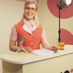 Nainen seisoo studiossa pöydän takana, edessään mikrofoni ja papereita.