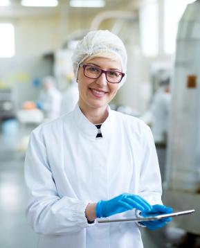 Nuori iloinen nainen, työntekijä steriileissä vaatteissa, joilla on tabletti ja hymyilee kameralle lähellä tehtaan tuotantolinjaa.